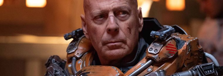 Bruce Willis from the shoulders up looks off to the viewer's right. He is wearing battered futuristic armor that is orange and silver.