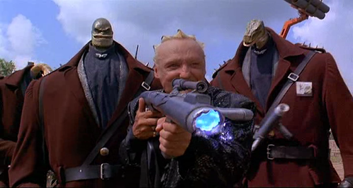 A still from the Super MArio Bros moive: Dennis Hopper, as King Koopa, shoots some sort of futuristic rocket launcher. He's flanked by large guys with tiny dinosaur/lizard heads.