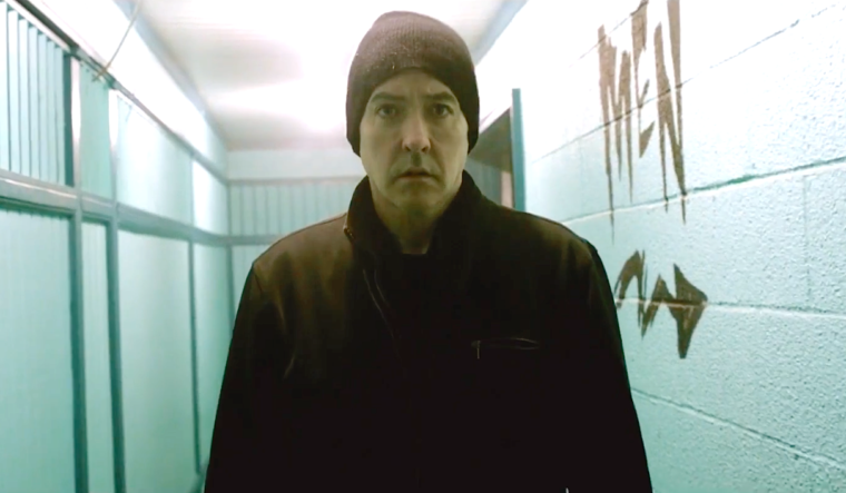 John Cusak, in a coat and cap, stands in a brightly lit hallway with what might be blood smeared on the walls. He's looking right of the camera.