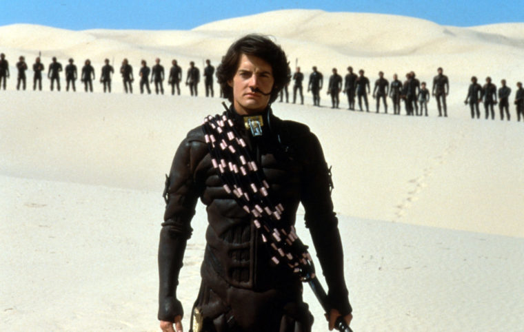 Kyle MacLachlan as Paul Mu'uad D'b stands wearing a stillsuit in the open desert. A row of figures stands very far behind him. He has a coil of rope over one shoulder, and is holding a staff in the opposite hand