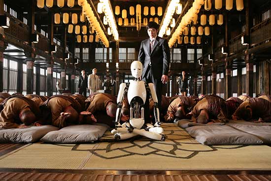 A robot with a white casing sits cross-legged in a room of monks all bowed down touching their heads to the floor. A man in a business suit stands behind the robot, angles and looking toward the robot. Prayer flags hang from the ceiling, and a stylized lotus flower in woven into the floor mat.