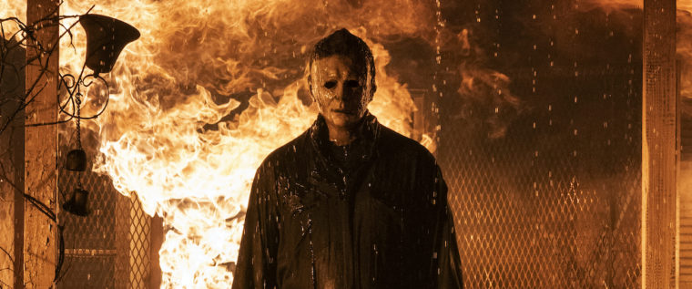 Michael Myers wearing his iconic white mask and black clothing stand on the porch of a building engulfed in flame