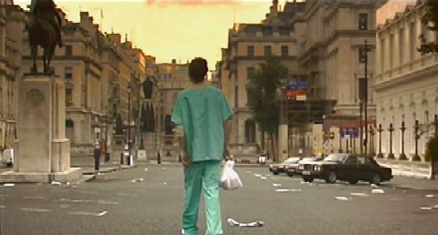 A still from 28 Days Later. A man in blue scrubs stands with his back to the viewer. In the background is a London streetscape empty of people and littered with trash.
